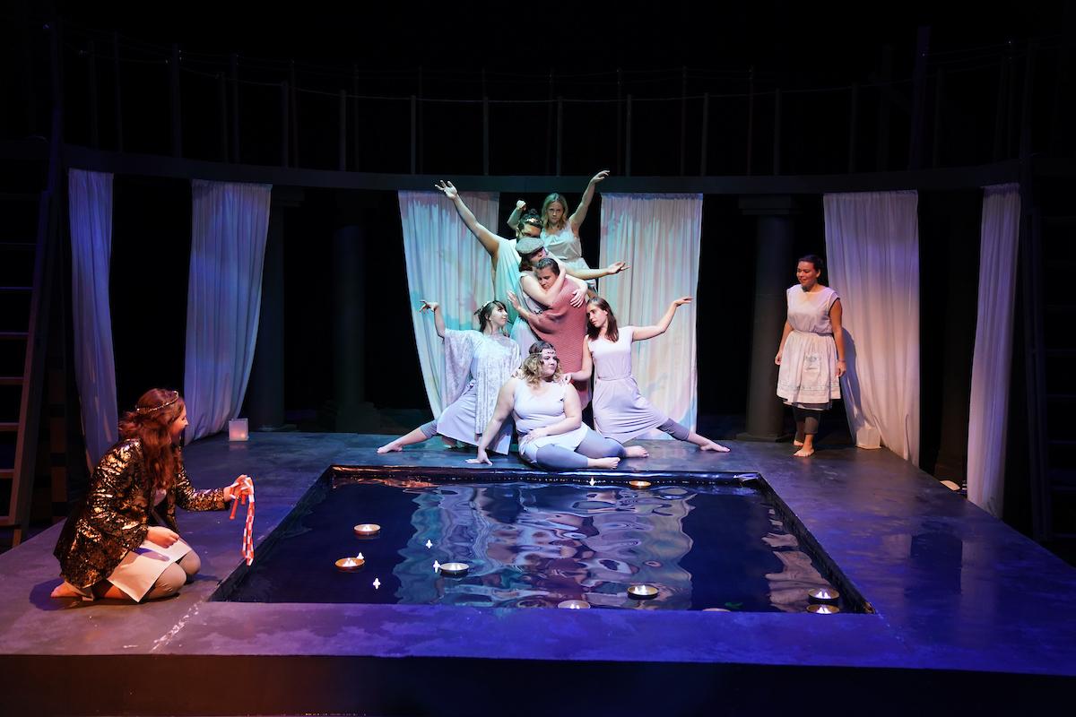 Actors in clothes that look like white togas pose dramatically at the end of a pool of water.